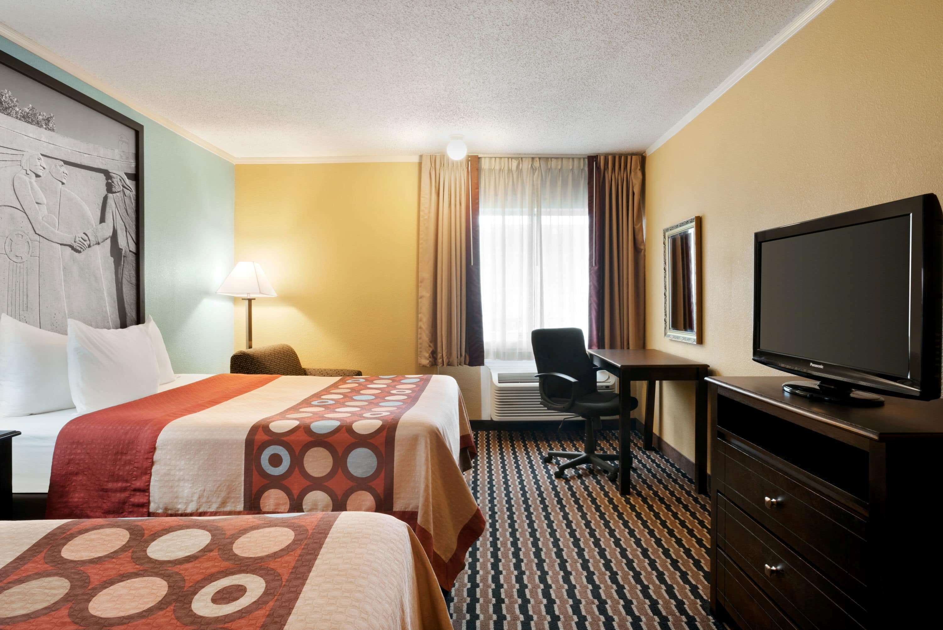 A hotel room with two beds, a desk, a television, and a large window with curtains.