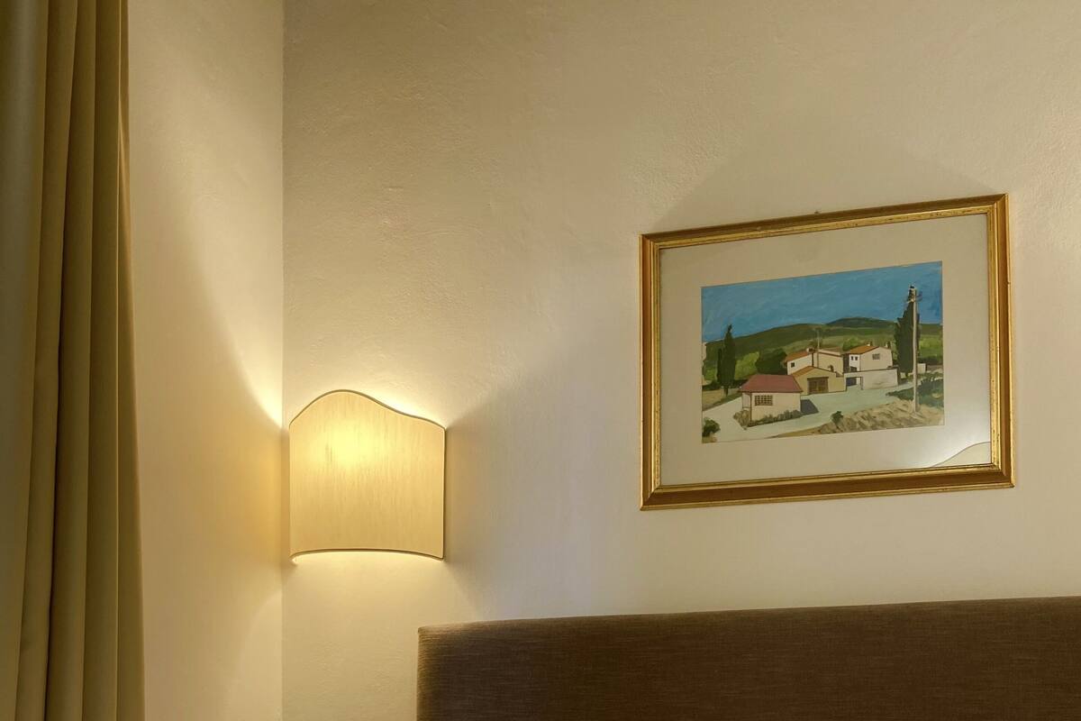 A neatly made bed with white linens and towels, a wall-mounted lamp, and a framed landscape painting.