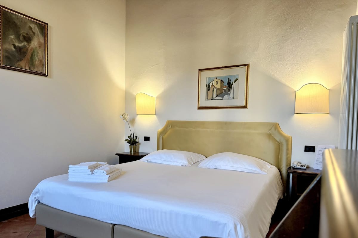 A hotel room with a bed, a nightstand with a lamp, a framed picture on the wall, and a small potted plant.
