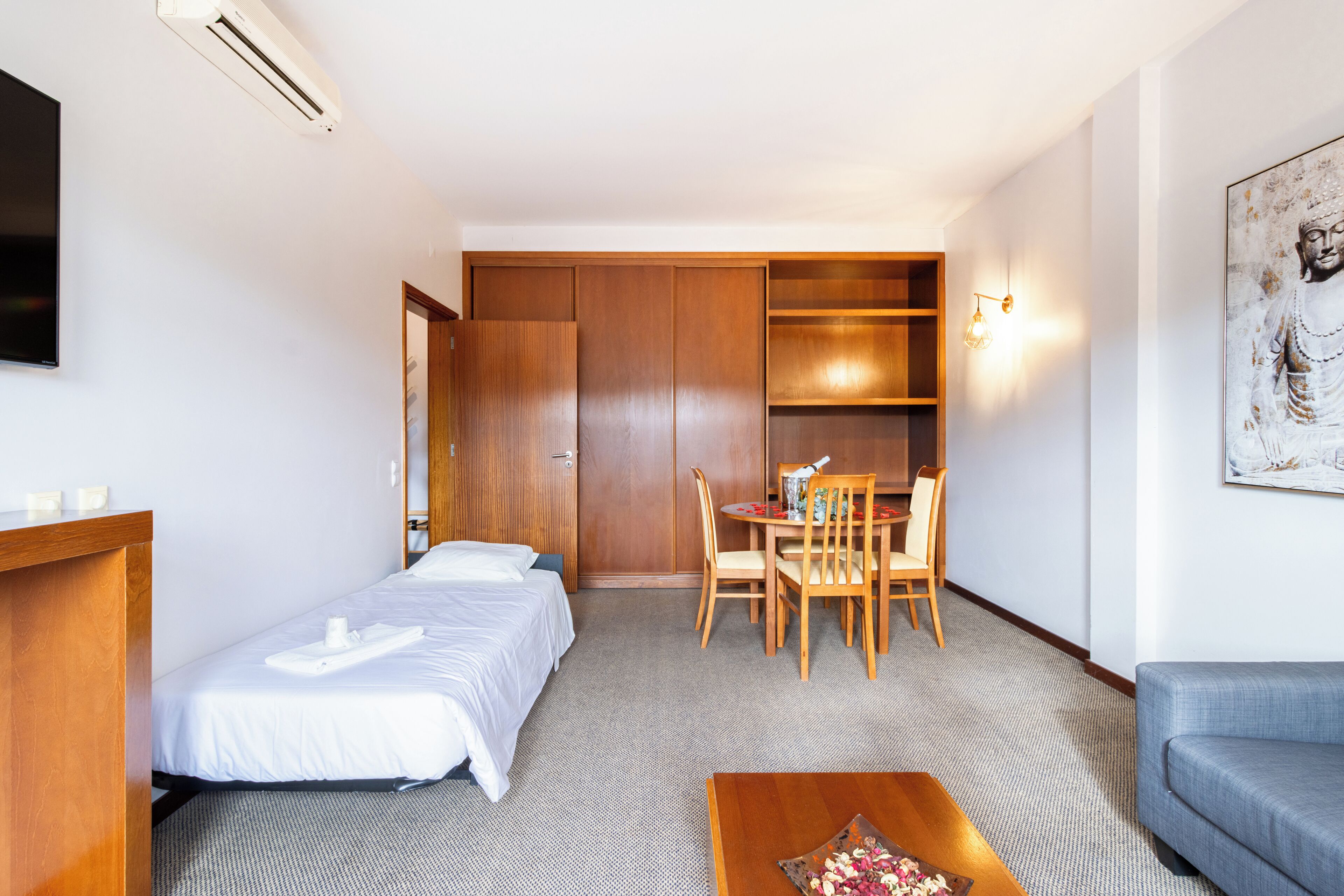 Una habitación de hotel con dos camas individuales, un escritorio de madera, una mesa de comedor con sillas, un sofá y un televisor montado en la pared.