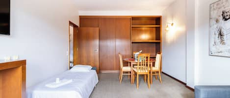 A hotel room with two single beds, a wooden desk, a dining table with chairs, a sofa, and a wall-mounted TV.