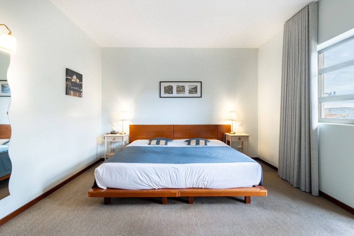 A hotel room with a large bed, two bedside tables, a headboard, and a window with curtains.