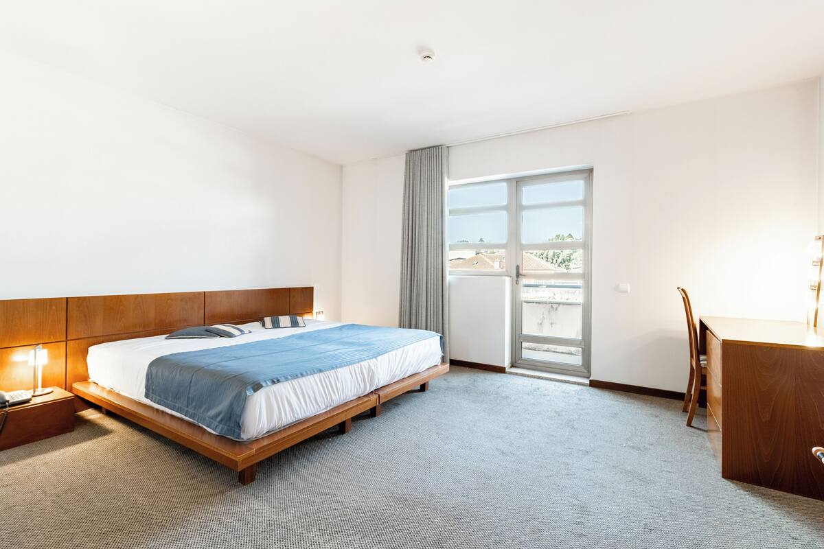 A hotel room with a bed, a desk, a chair, a nightstand, and a window with a view.
