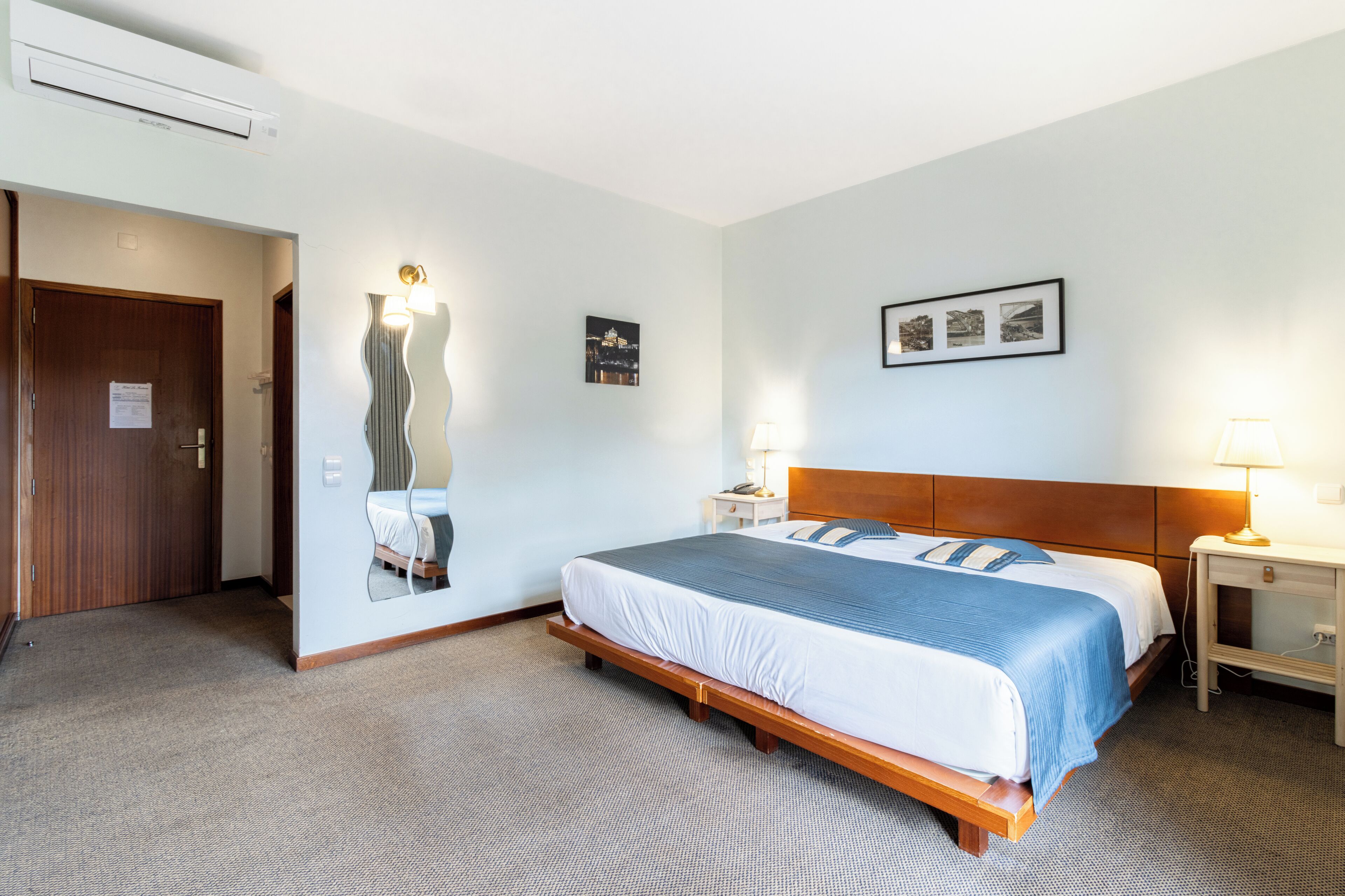 Une chambre d’hôtel avec un grand lit, une tête de lit en bois, deux tables de chevet avec des lampes, un miroir fixé au mur et une porte.
