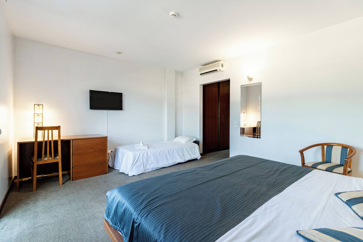 A hotel room with a large bed, a smaller bed, a desk, a chair, a TV, and a wardrobe.