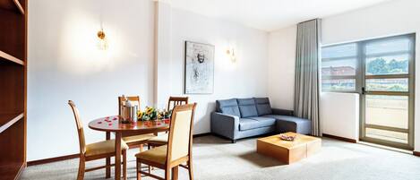 A modern hotel room with a dining area, a sofa, and a large window offering a view.
