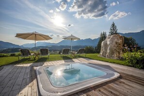 Un jacuzzi en una terraza de madera con dos sombrillas, rodeado de sillas y una gran roca, con vistas a montañas.