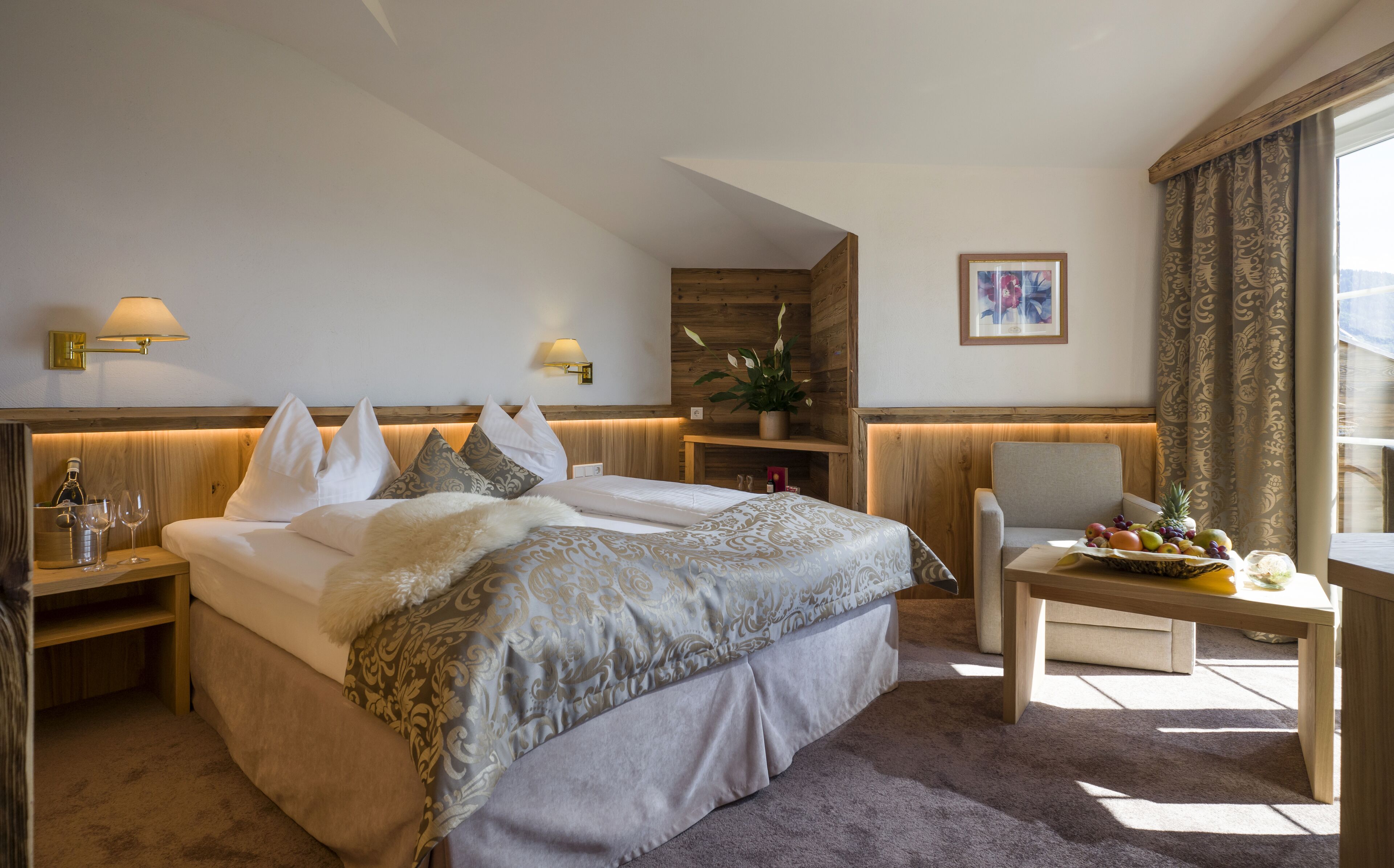 A hotel room with a large bed, a chair, a small table with fruit, a plant, and a painting on the wall.