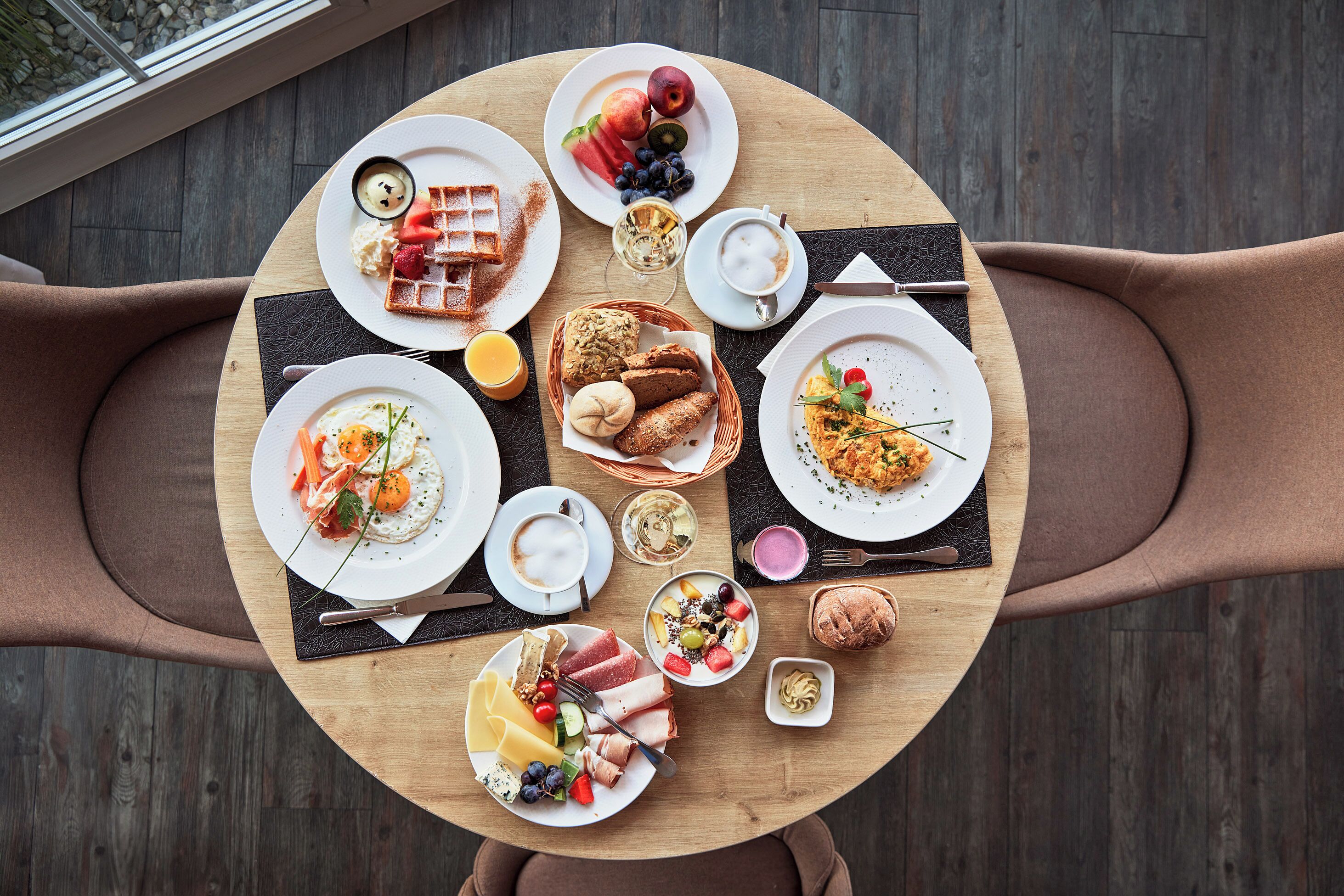Una mesa de madera redonda con alimentos para el desayuno, como tostadas, fruta, huevos y pasteles, rodeada de sillas marrones.