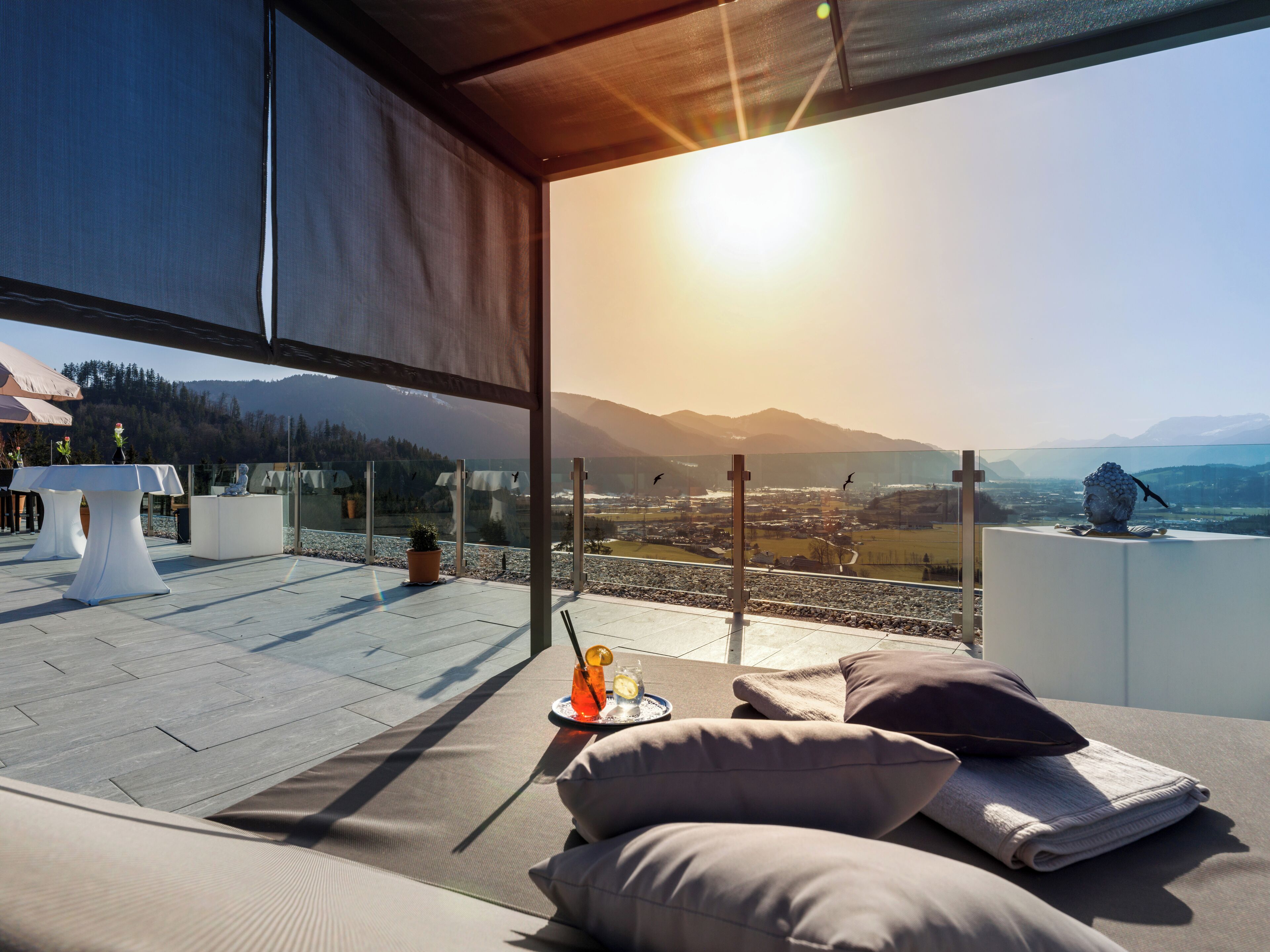 A rooftop terrace with a clear view of a valley and mountains, furnished with cushions and a table with drinks.