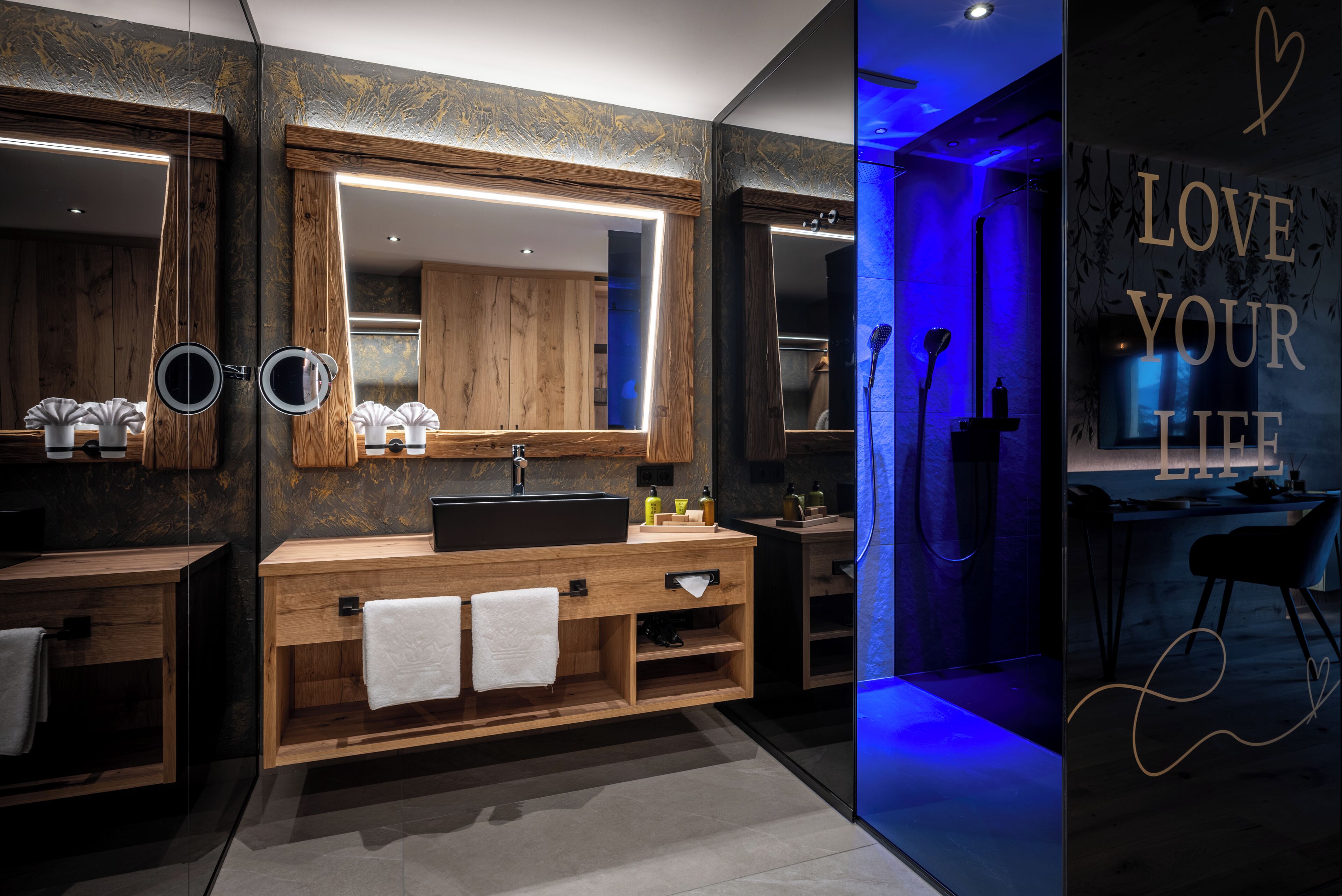 A modern bathroom with wooden vanity, large mirrors, and a blue-lit wall with a motivational quote.
