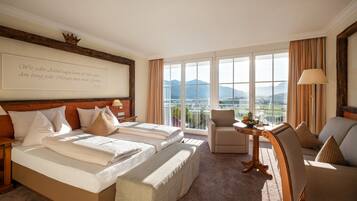 A hotel room with two beds, a sofa, a chair, a small table with fruit, and a large window with a mountain view.