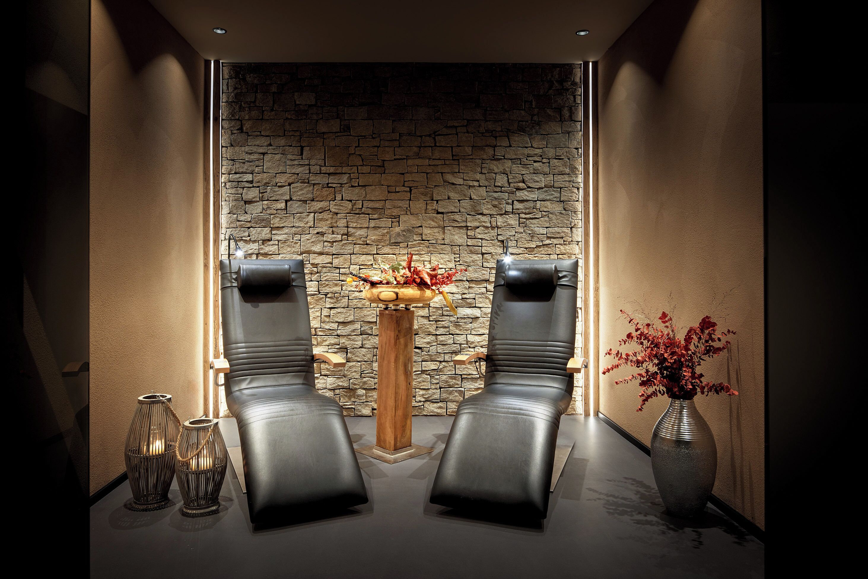 Un espace spa comprenant deux fauteuils en cuir noir, un mur en pierre, une table en bois ornée d’un vase de fleurs et deux vases décoratifs.