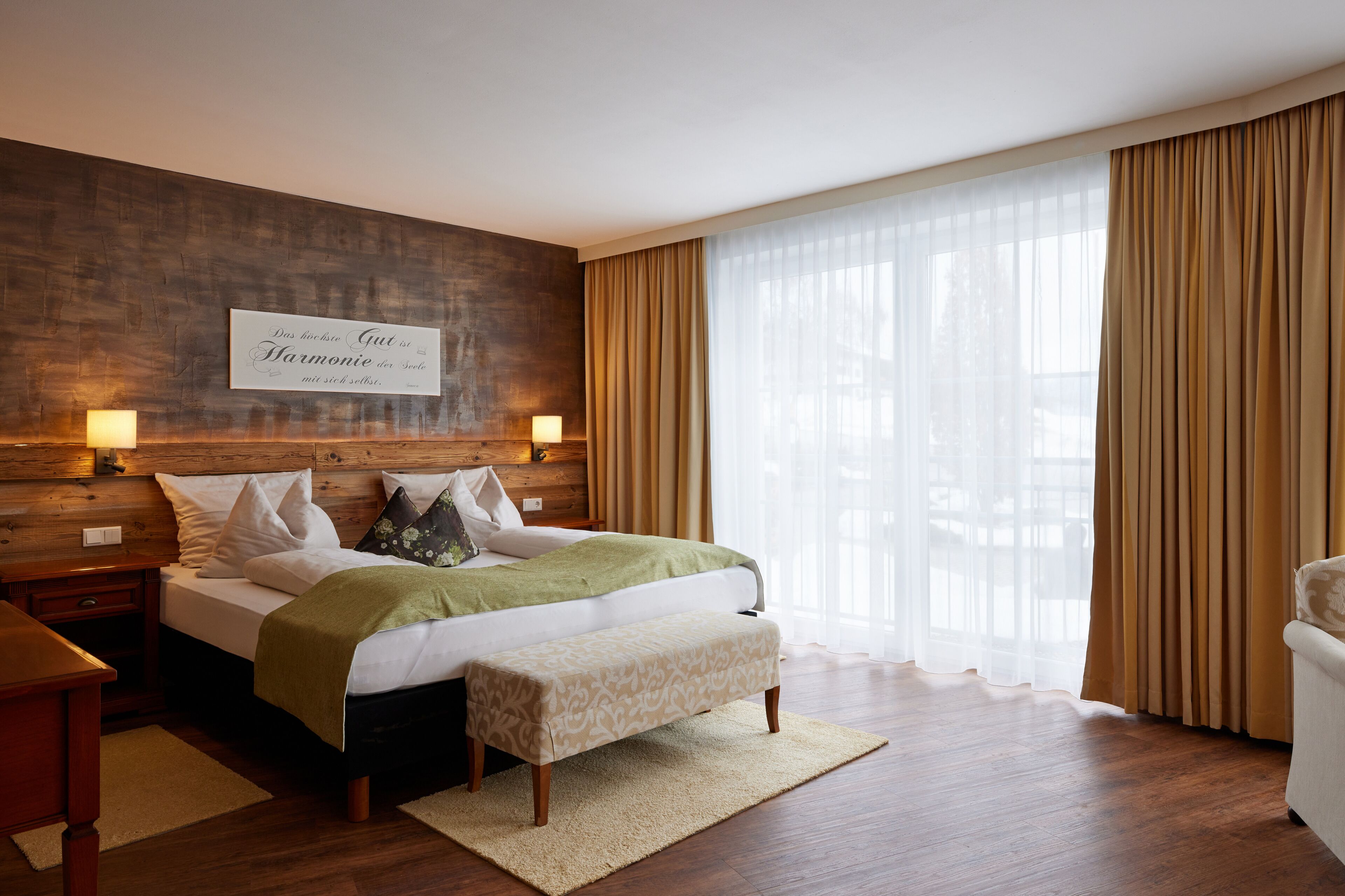 Een moderne hotelkamer met een houten hoofdeinde, een groot bed met een groene plaid, een bankje en een groot raam met dunne gordijnen.