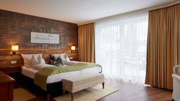 Una habitación de hotel moderna con cabecera de madera, una cama grande con una manta verde, un banco y un ventanal con cortinas translúcidas.