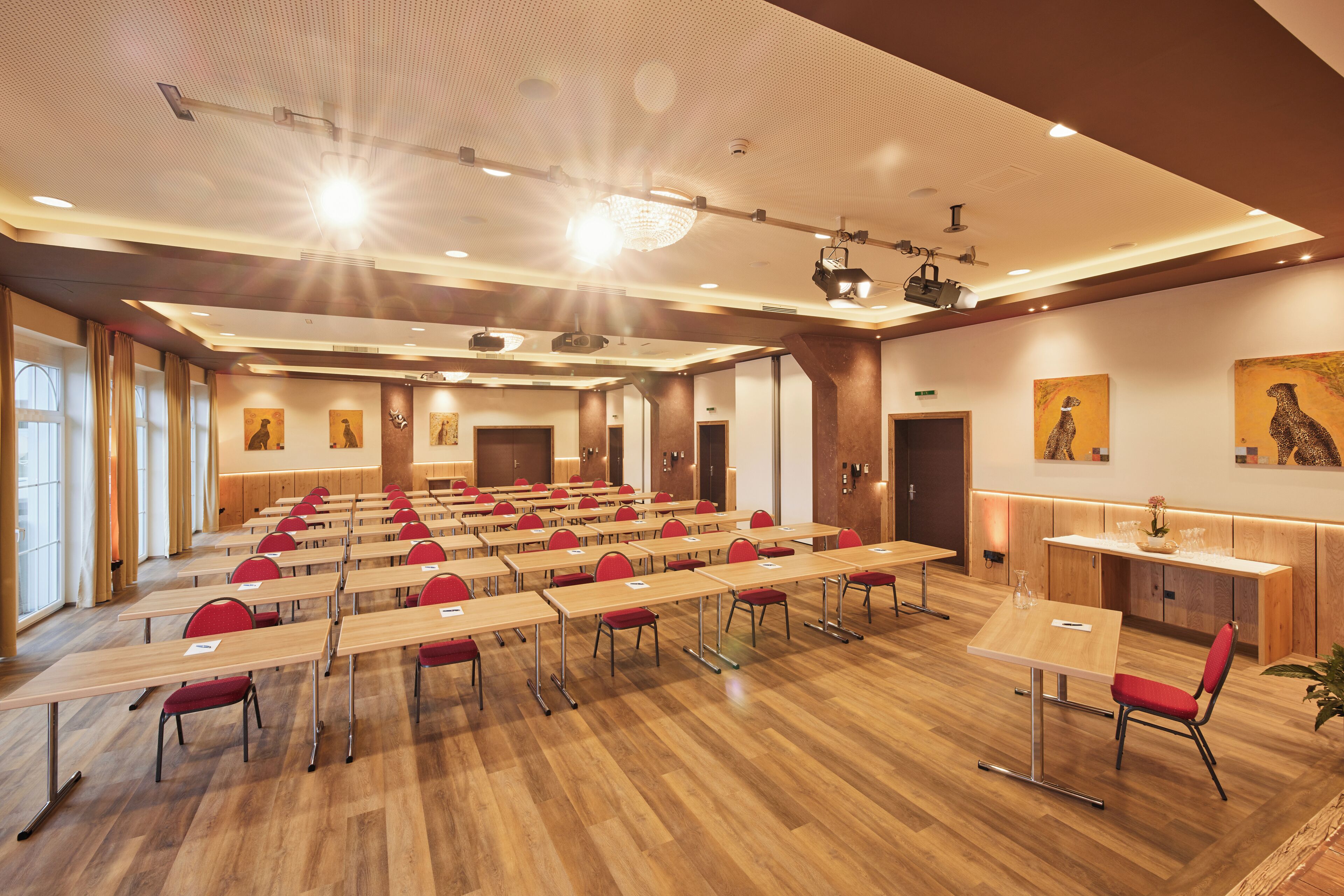 Un salón de conferencias con filas de mesas y sillas, un escenario con proyector y obras de arte en las paredes.