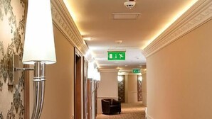 A hotel corridor with patterned carpet, wall sconces, and directional signs. - Hotel Capitulum (Gyor)