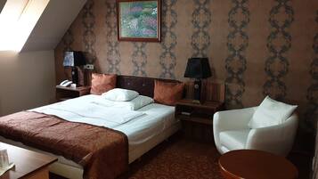 Une chambre d’hôtel avec un lit, deux tables de chevet avec des lampes, un fauteuil blanc, une petite table ronde et un tableau au mur.