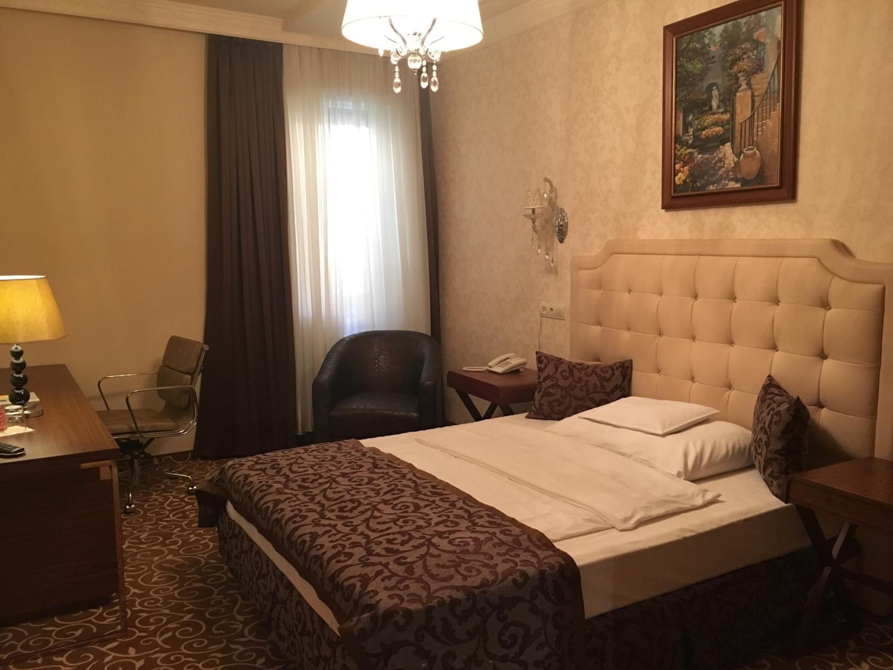 A hotel room with a large bed, a desk with a lamp, a chair, a telephone, and a painting on the wall.
