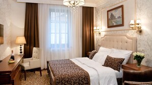 A hotel room with a large bed, a desk with a lamp, a chair, and a nightstand with a vase of flowers. - Hotel Capitulum (Gyor)