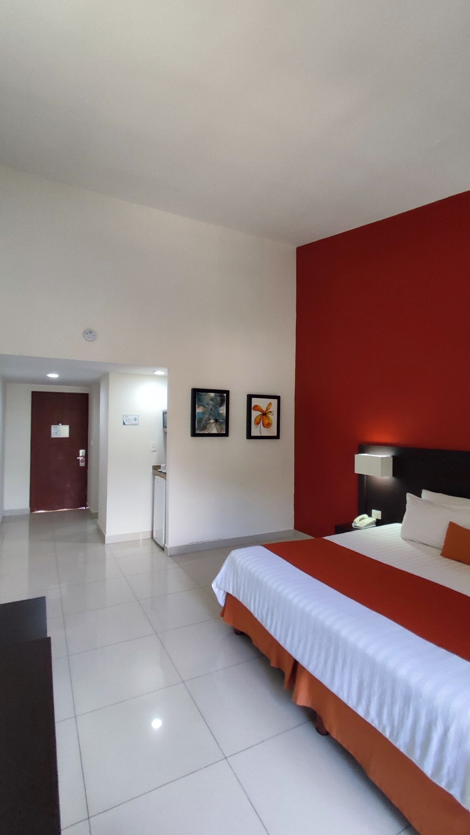 A hotel room with a large bed, a red accent wall, and two framed pictures on the wall.