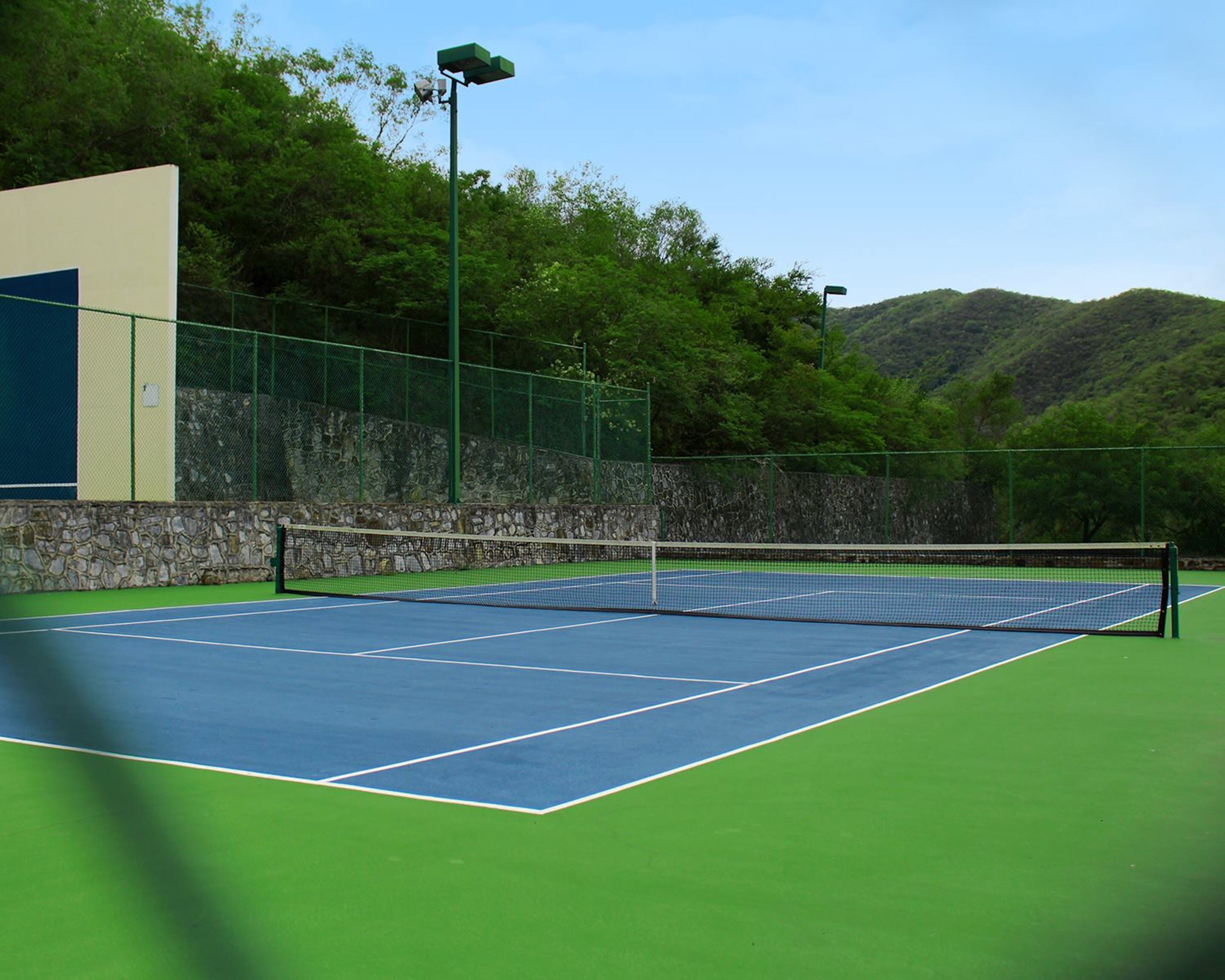 En udendørs tennisbane med net, omgivet af en stenvæg og grøn hegn.