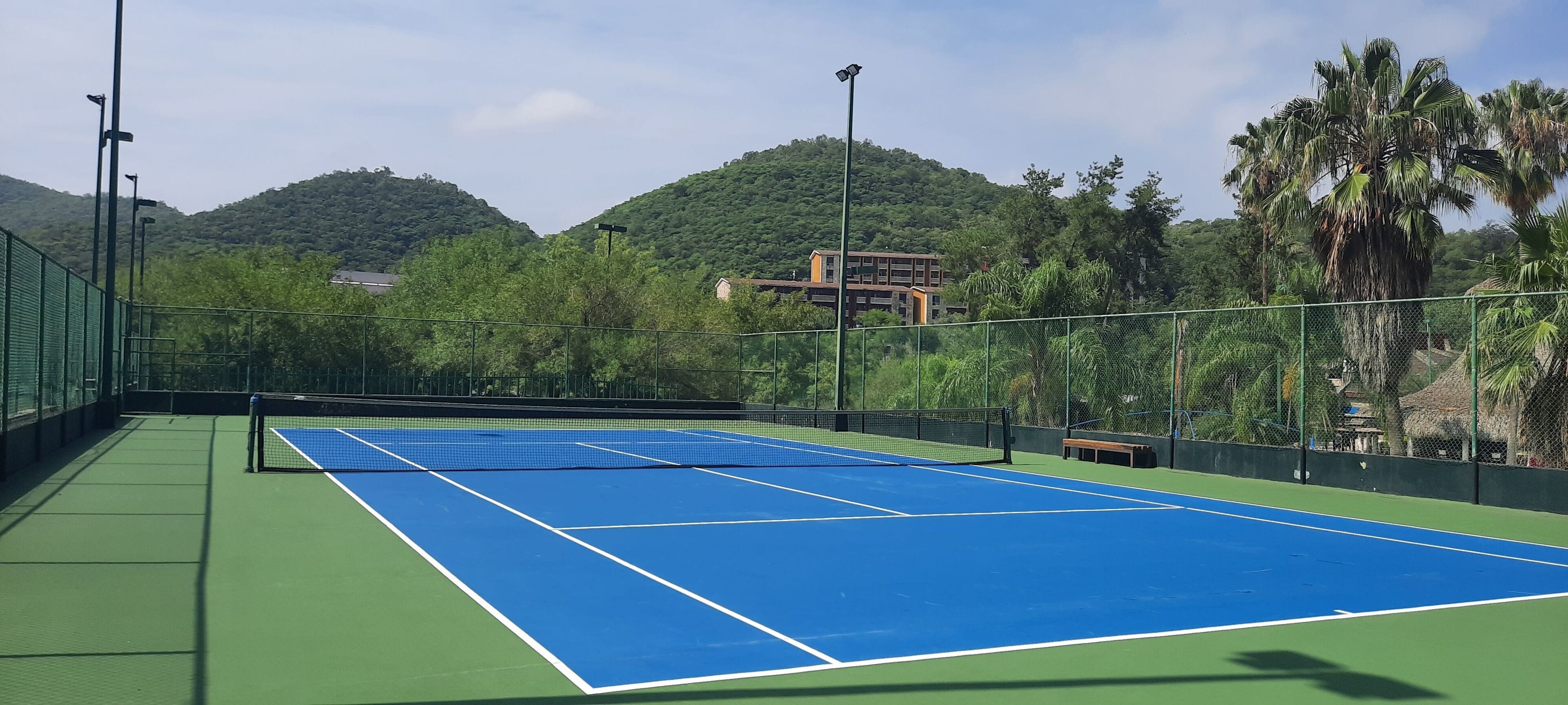 An outdoor tennis court with a net, surrounded by a green fence and trees.