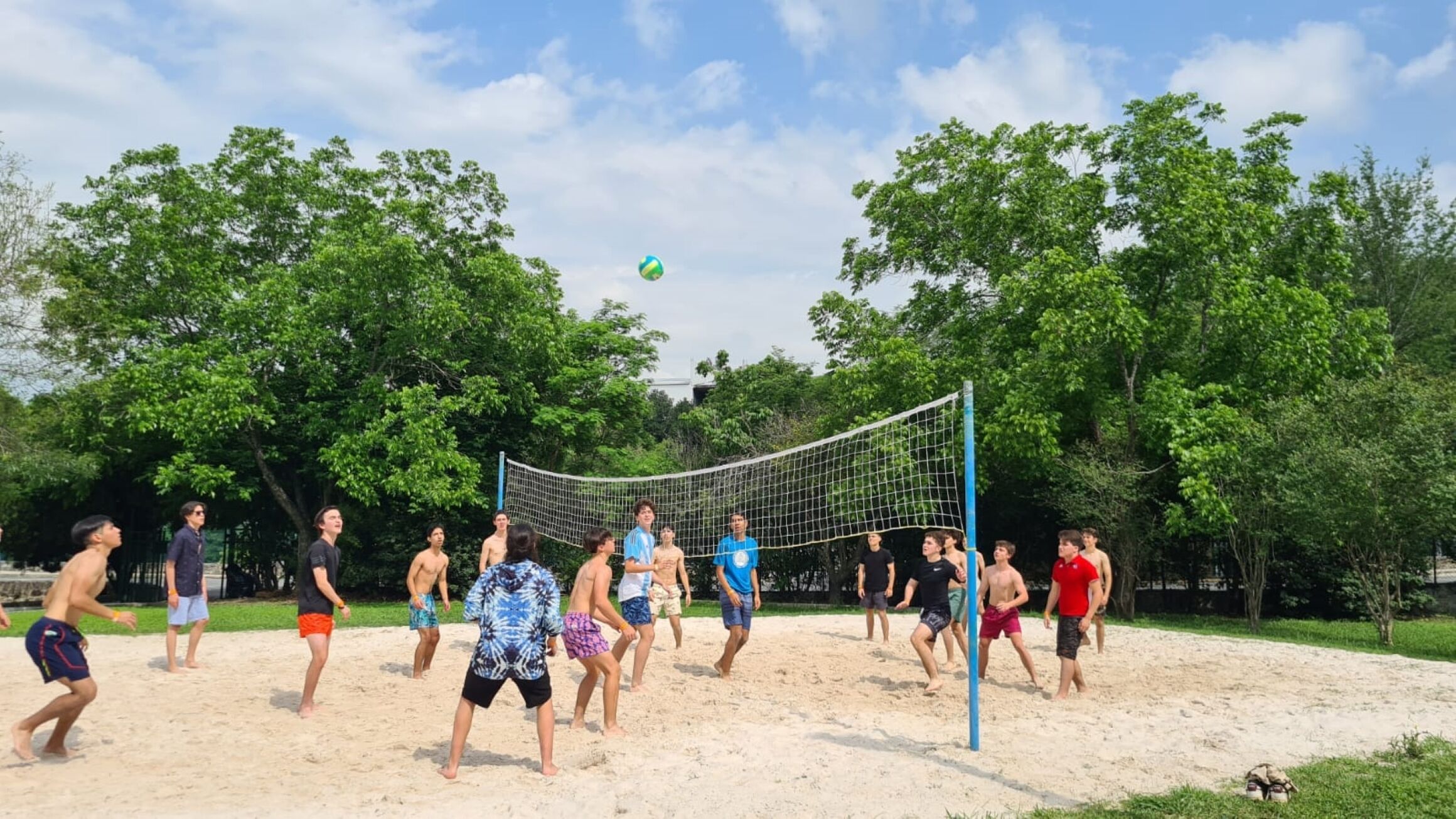 Un grupo de personas juega voleibol de playa en una cancha de arena rodeada de árboles.