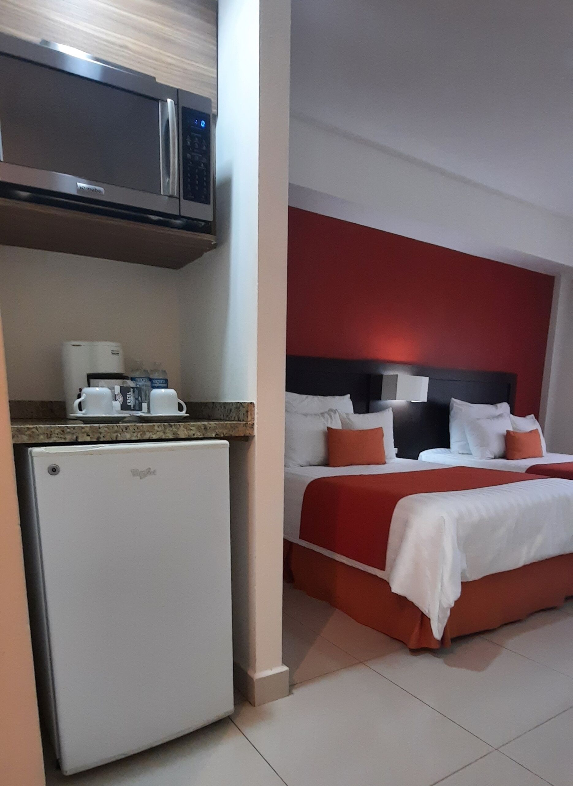 A compact hotel room with a kitchenette, a bed with red and white bedding, and a modern headboard.
