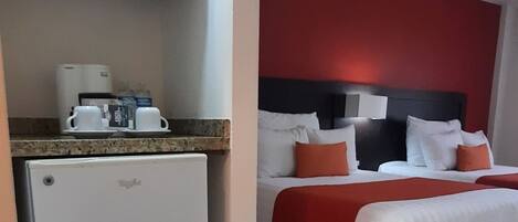 A compact hotel room with a kitchenette, a bed with red and white bedding, and a modern headboard.