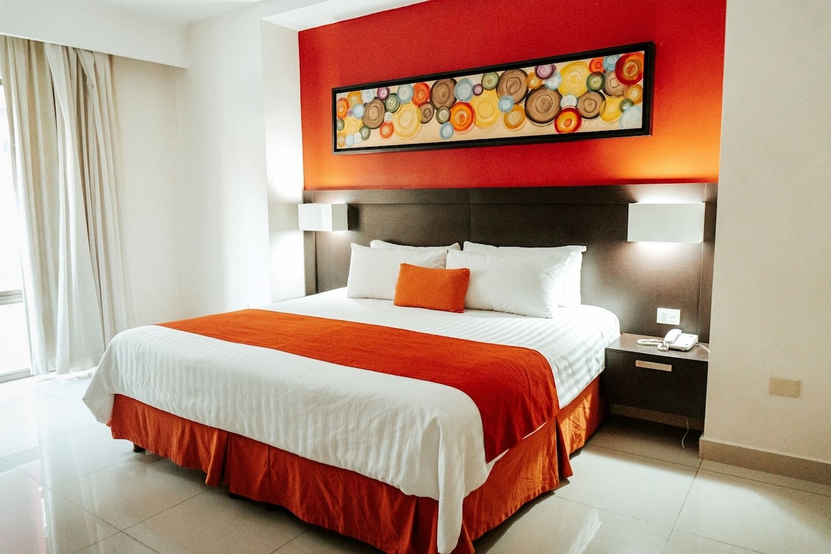 Habitación de hotel con una cama grande, ropa de cama blanca, una pared de acento roja, un cuadro sobre la cama y una mesita de noche con un teléfono.