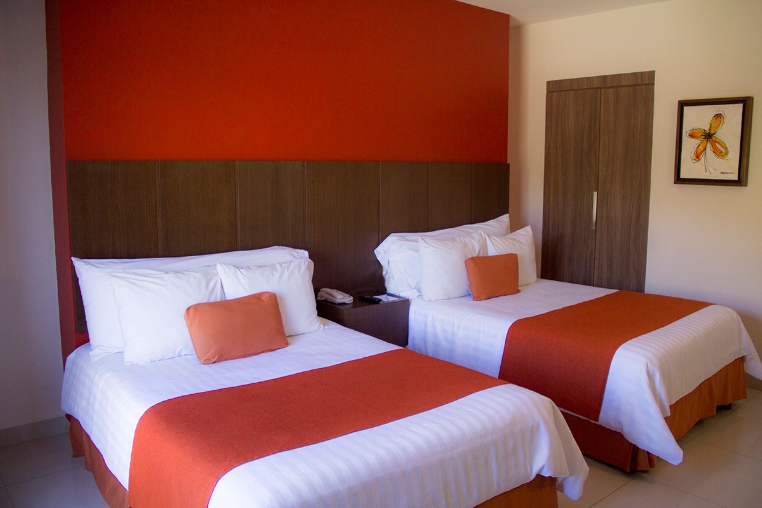 Two double beds with white and orange bedding, a wooden headboard, a wooden wardrobe, and a framed artwork on the wall.