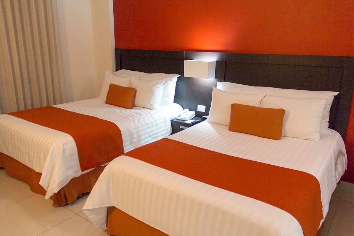 Habitación de hotel con dos camas, cada una con ropa blanca y detalles anaranjados, una mesita de noche con lámpara y una pared de acento roja.