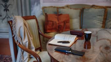 A round wooden table with a notebook, pen, and a cup. A chair with a blanket and a handbag is visible.