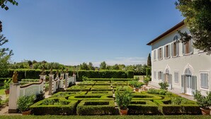 Un jardín formal con setos geométricos, un edificio clásico con ventanas arqueadas y estatuas.