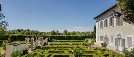 A formal garden with geometric hedges, a classical building with arched windows, and statues.