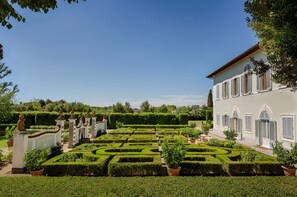 Un jardín formal con setos geométricos, un edificio clásico con ventanas arqueadas y estatuas.