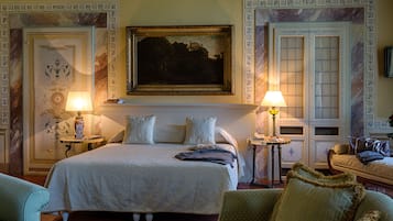 Una habitación de hotel elegante con una cama grande, dos mesitas de noche, un cuadro sobre la cama y una zona de estar con un sofá y sillones.