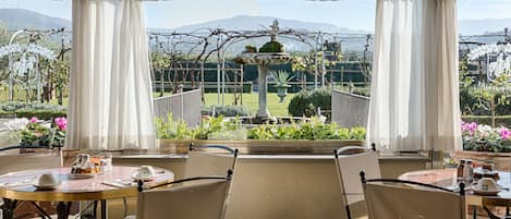 A conservatory with a table set for two, surrounded by chairs, overlooking a garden with a fountain and a mountain in the background.