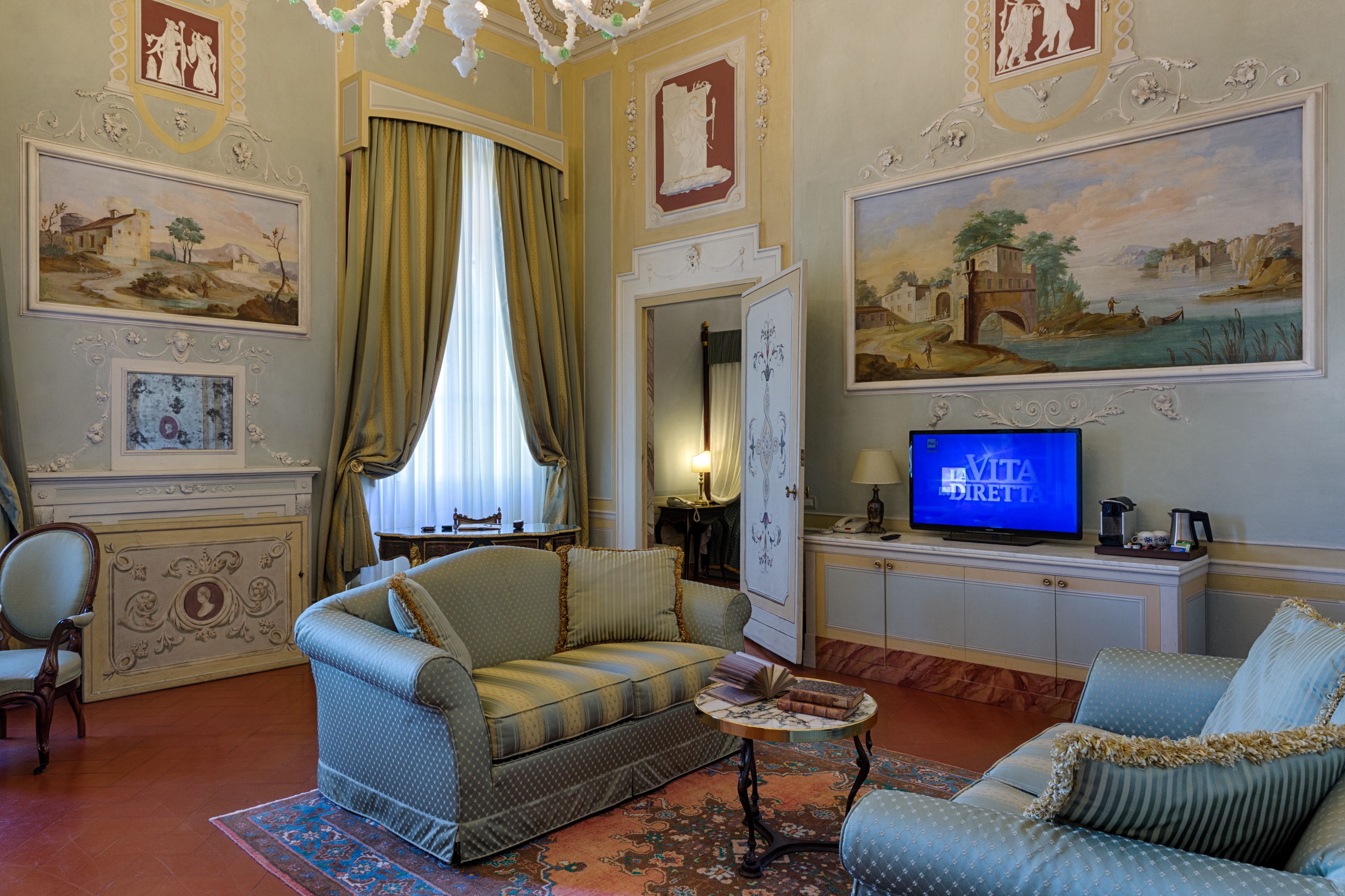 A room with a sofa, armchair, and a television. There are paintings on the walls and a window with curtains.