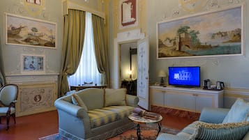 A room with a sofa, armchair, and a television. There are paintings on the walls and a window with curtains.