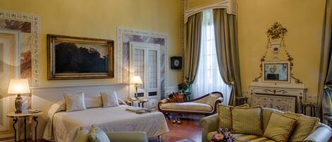 A hotel room with a large bed, two armchairs, a sofa, a small table with a vase of flowers, and a fireplace.