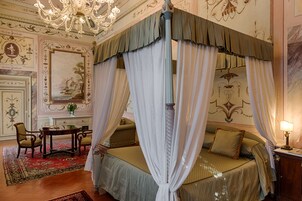 Un letto a baldacchino con tende verdi, drappeggi bianchi, un lampadario, decorazioni murali elaborate e un tavolino con sedie in una stanza con tappeto rosso.