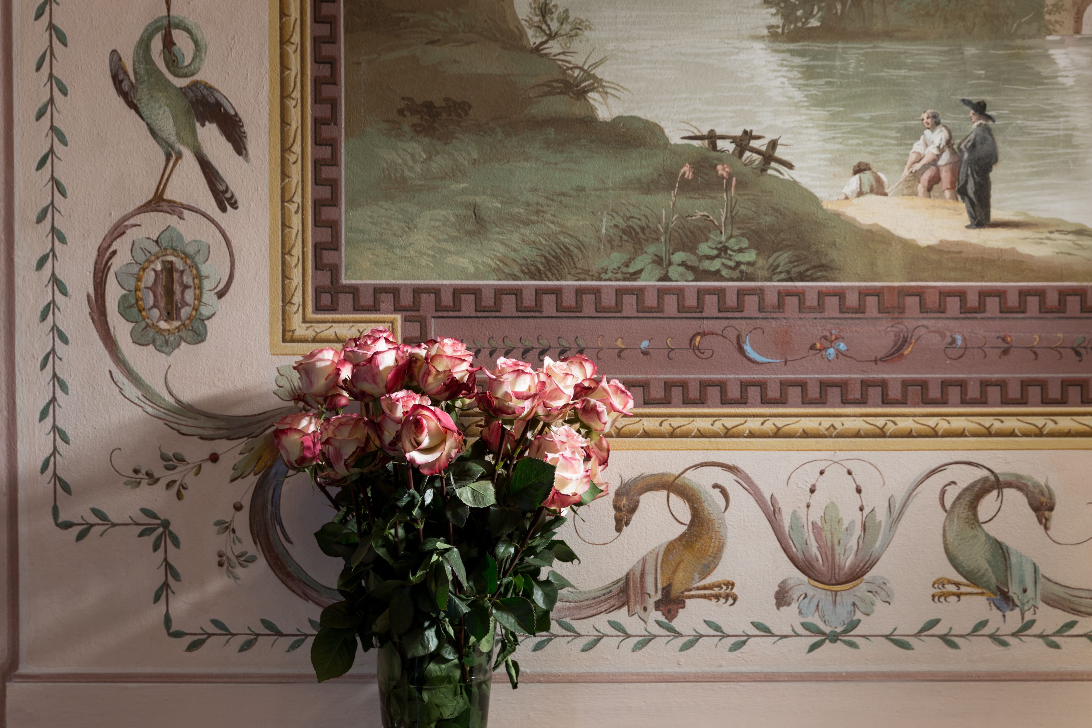 Een kamer met een muurschildering van een vredig landschap, een vaas met roze rozen en decoratieve muurpatronen.