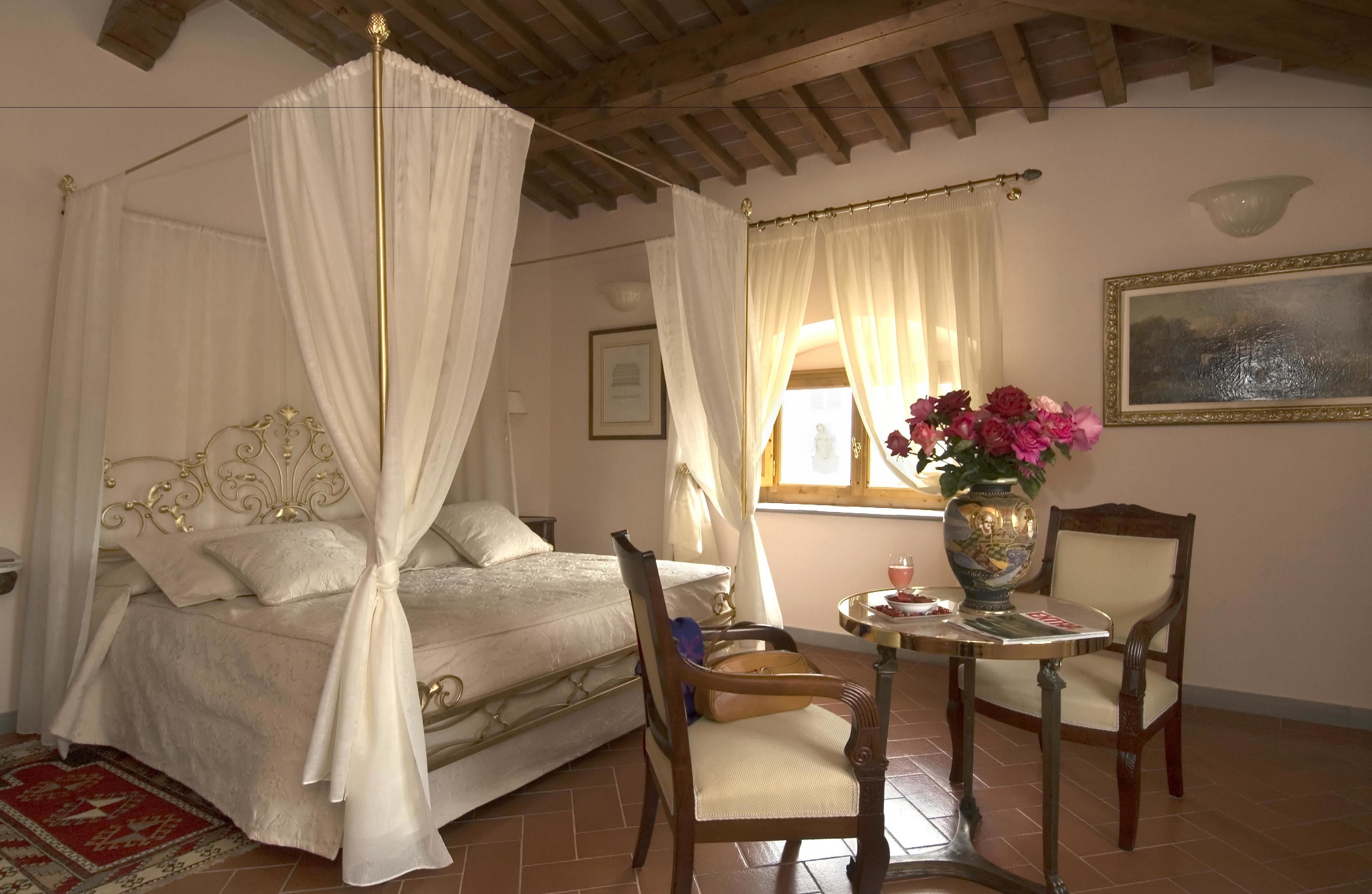 Un dormitorio con cama con dosel, una silla de madera, una mesita con un florero y un cuadro en la pared.