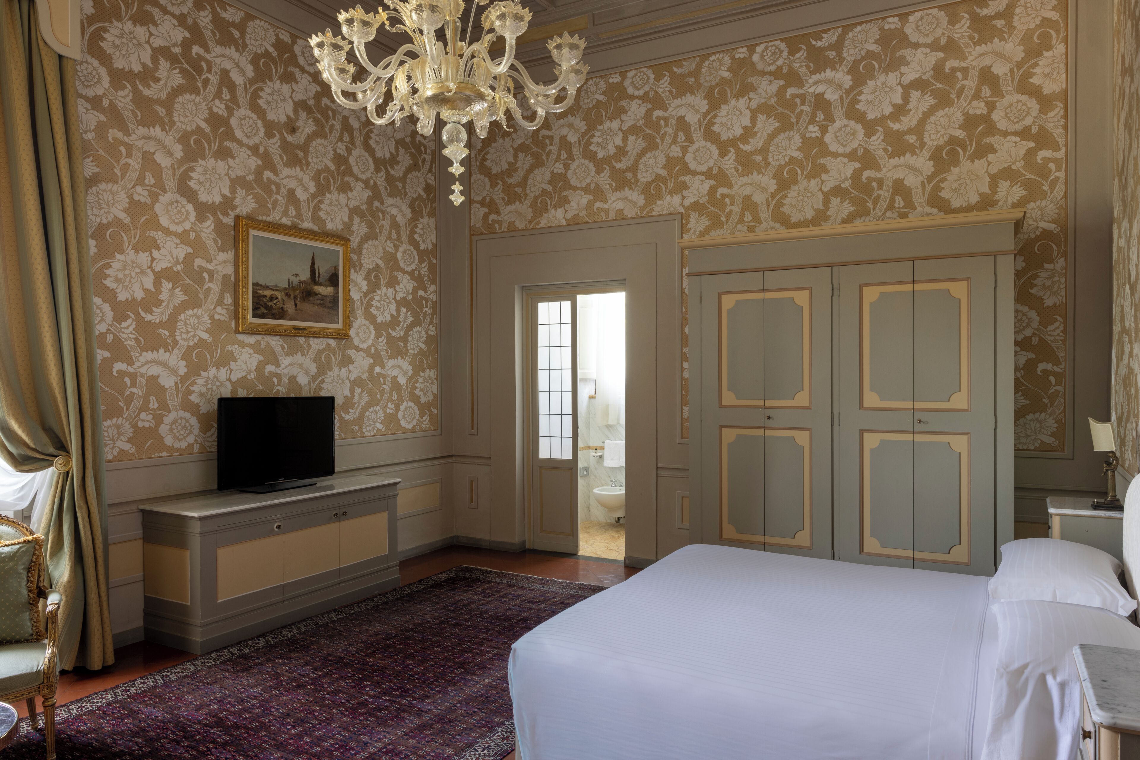 Ein Hotelzimmer mit einem Bett, einem Fernseher auf einer Kommode, einer Chandelüster, einem Kleiderschrank und einer Tür zu einem Badezimmer.