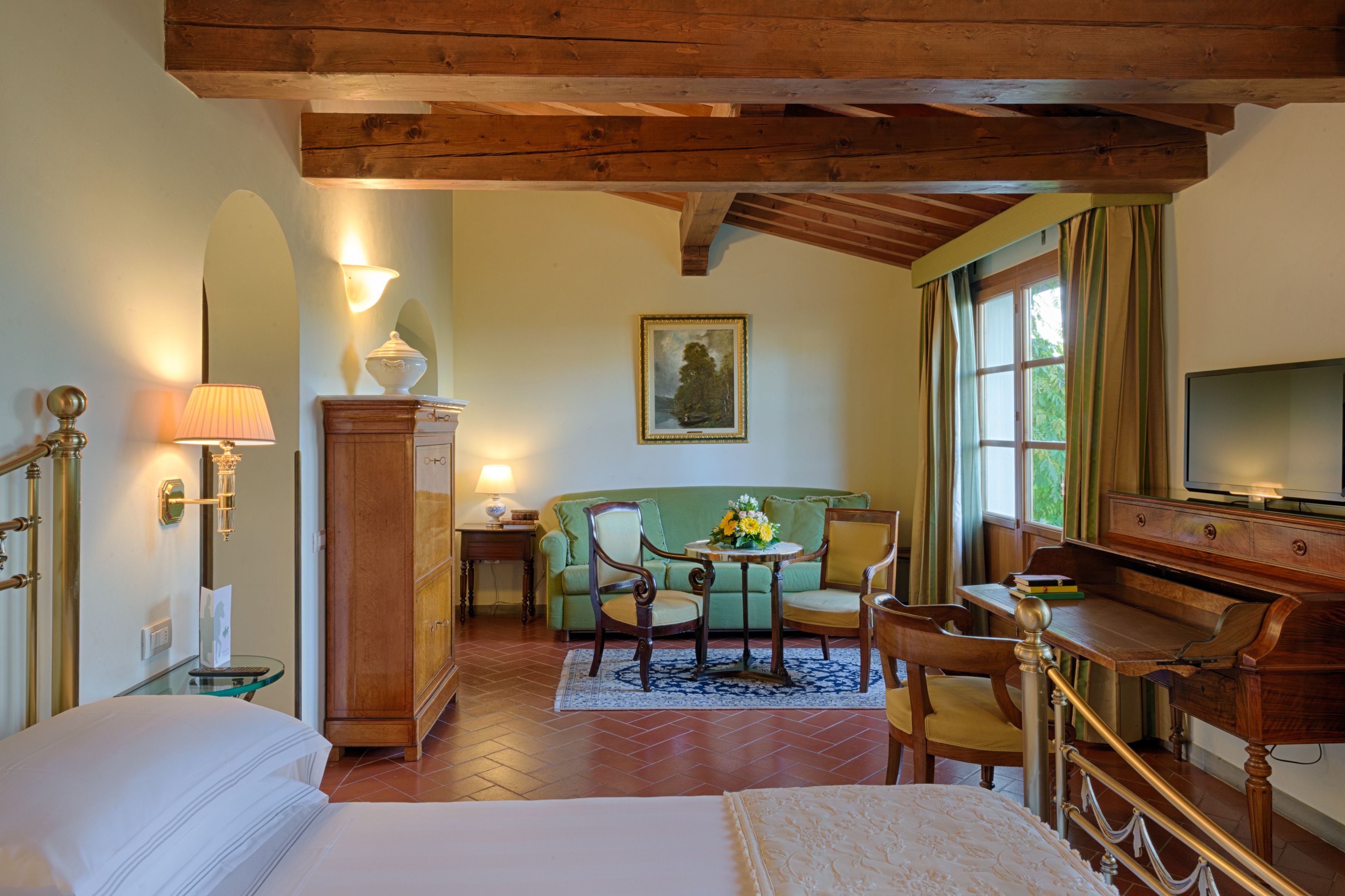 A hotel room with a bed, a wooden desk, a green sofa, a small table with flowers, and a painting on the wall.