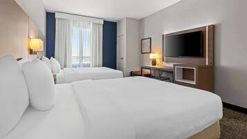 A hotel room with two beds, a desk with a chair, a flat-screen TV, and a window with curtains.