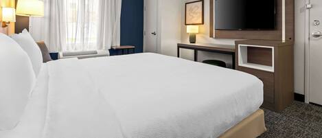 A hotel room with a large bed, a desk with a flat-screen TV, a window with curtains, and a door.