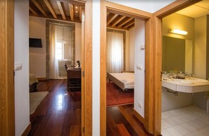 Une pièce avec un plancher en bois, un lit, un bureau et une salle de bain équipée d’un lavabo et d’un miroir.
